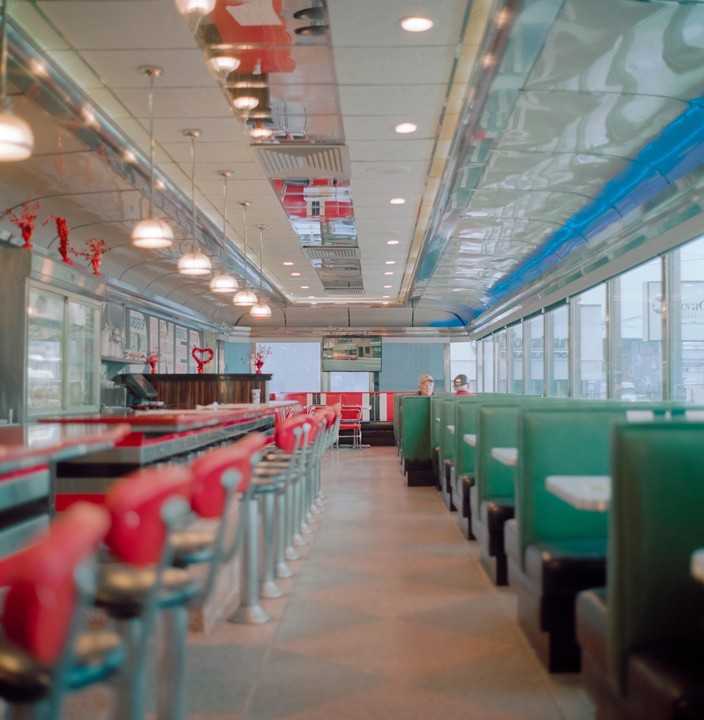 A diner interior features a long counter with red stools on the left and green booths on the right, with a man visible in one of the booths.