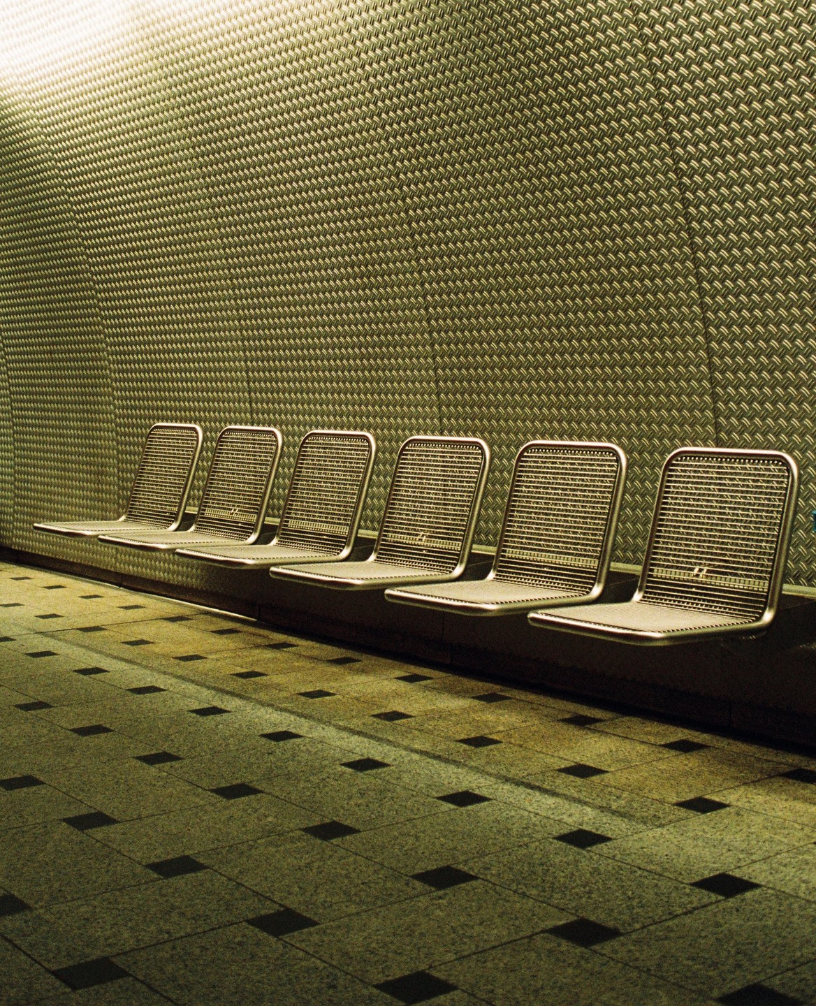 A row of empty metal chairs sits against a textured wall with a tiled floor in the foreground.
