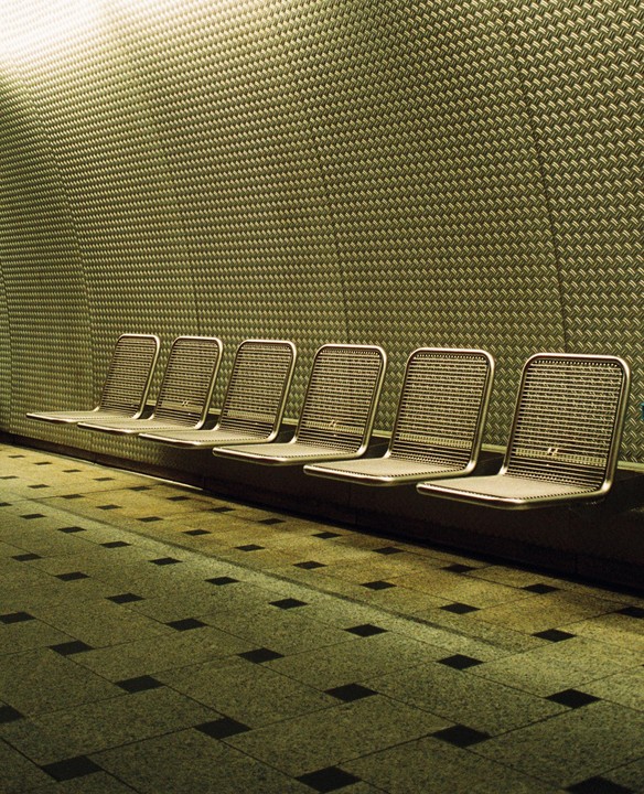 A row of empty metal chairs sits against a textured wall with a tiled floor in the foreground.