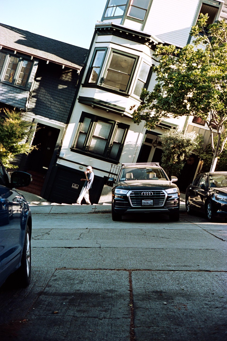A man walks on a steep street with parked cars and houses on either side.