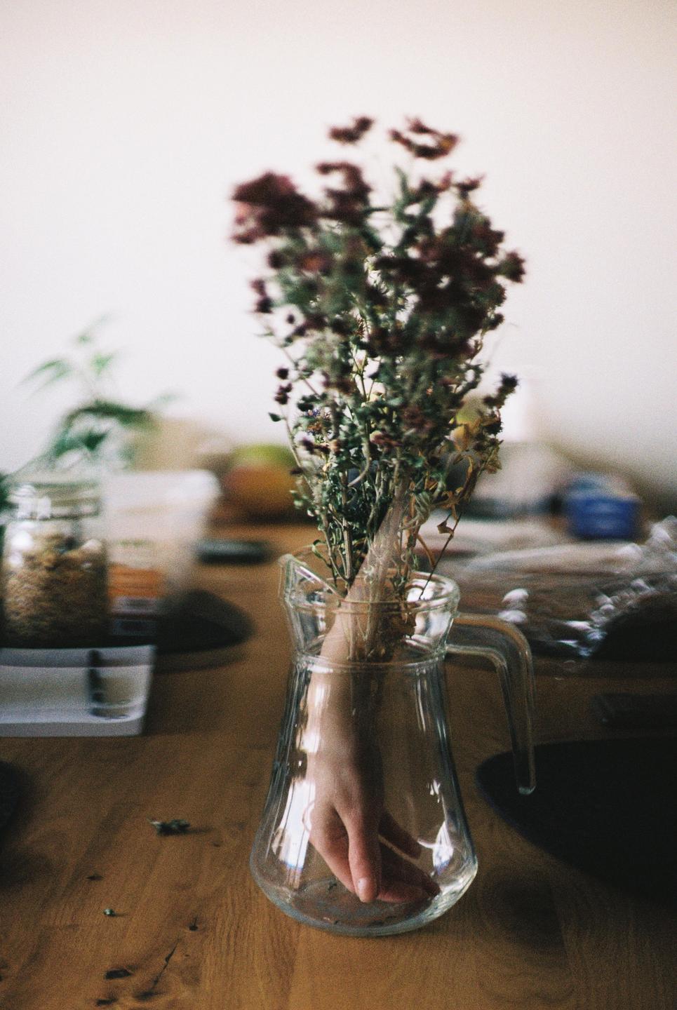 A hand is visible inside a glass pitcher holding a bouquet of dried flowers on a wooden table.
