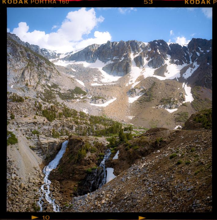 A waterfall cascades down a rocky mountain with snow-capped peaks under a blue sky.
