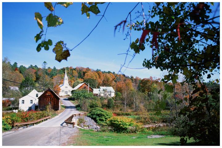A small town with a white church and several houses is nestled among hills covered in autumn foliage, with a road and bridge in the foreground.