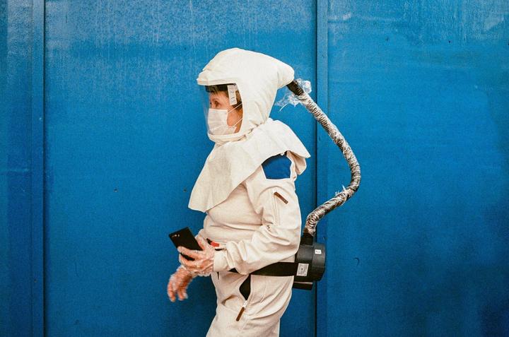 A person in a white protective suit and mask holds a phone against a blue wall.