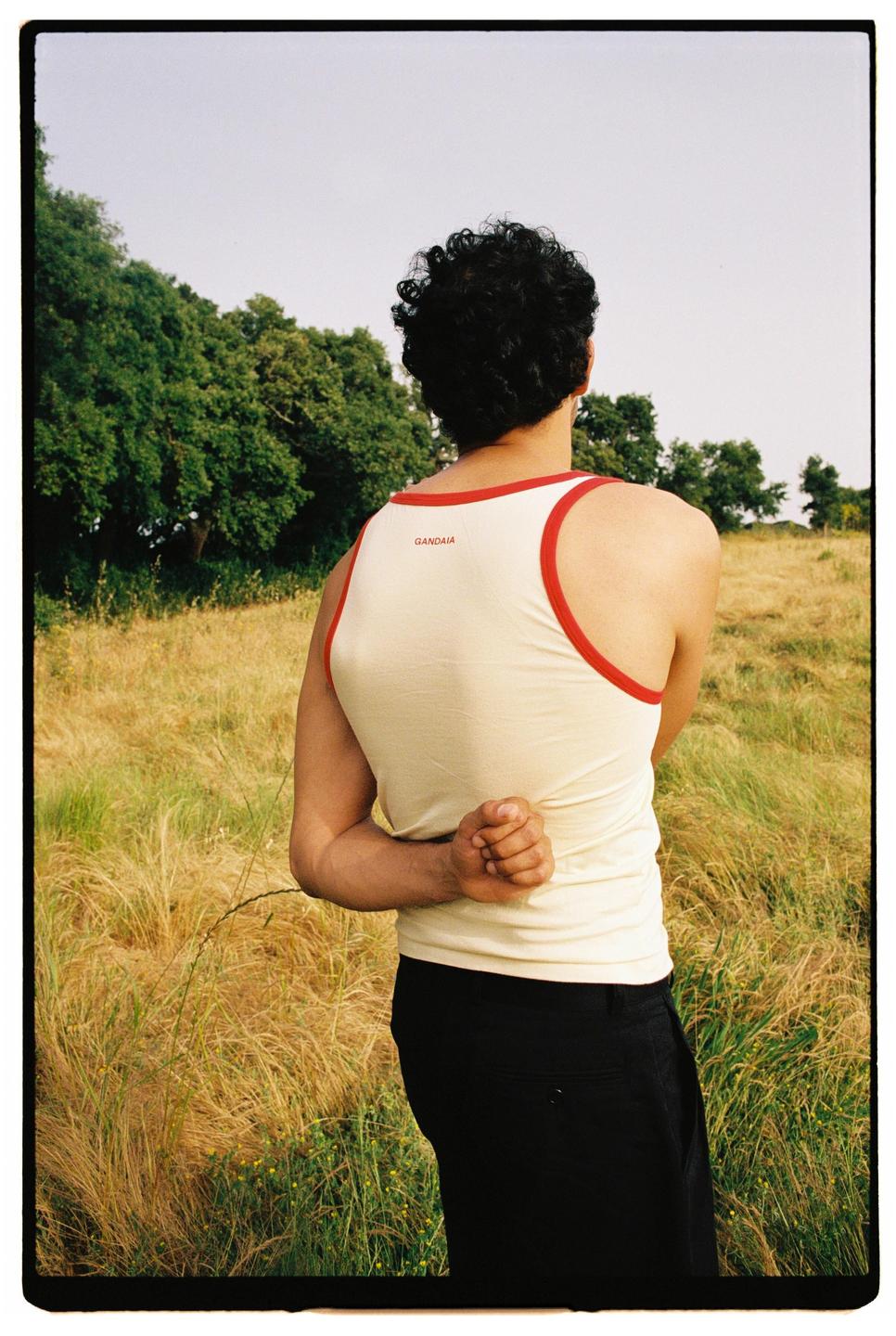 A man with curly hair stands in a field with his hands clasped behind his back, looking out at trees in the distance.