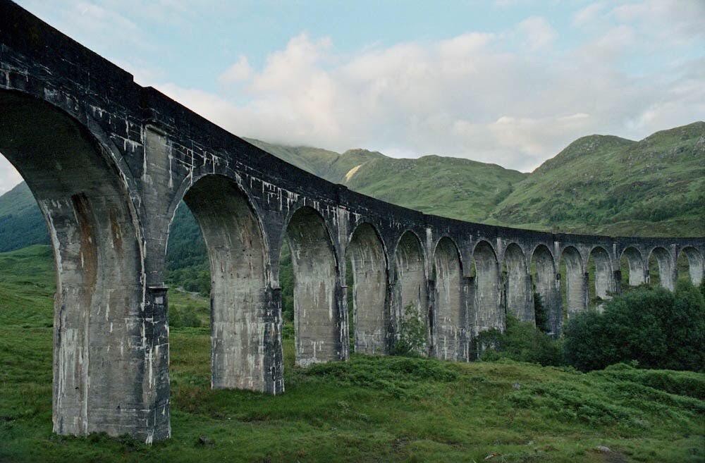 A long, arched viaduct stretches across a green valley with mountains in the background under a cloudy sky.