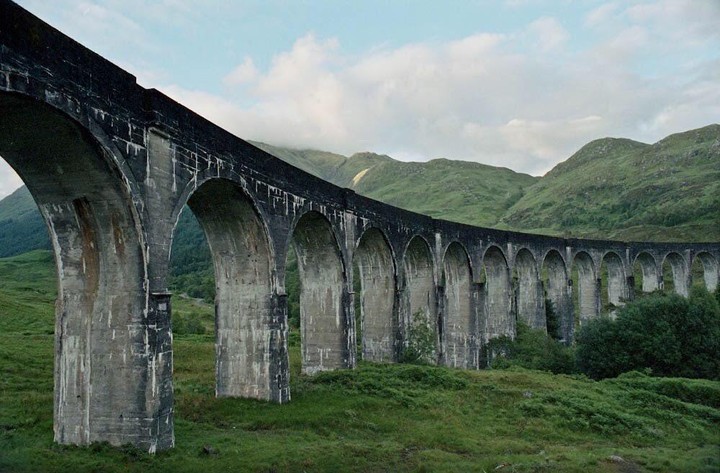 A long, arched viaduct stretches across a green valley with mountains in the background under a cloudy sky.
