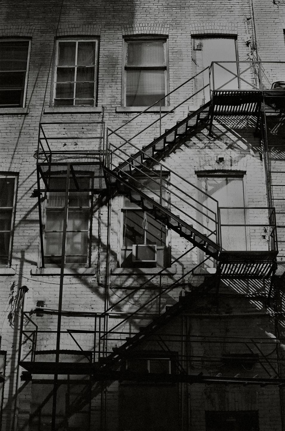 A fire escape with diagonal shadows is attached to a brick building with multiple windows.