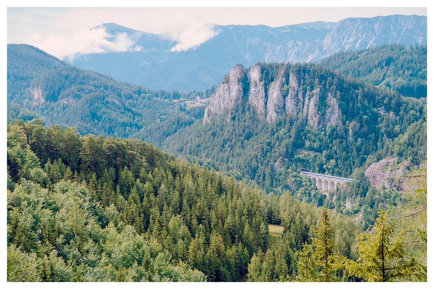 A blue train crosses a stone viaduct in a valley surrounded by mountains and dense forest.