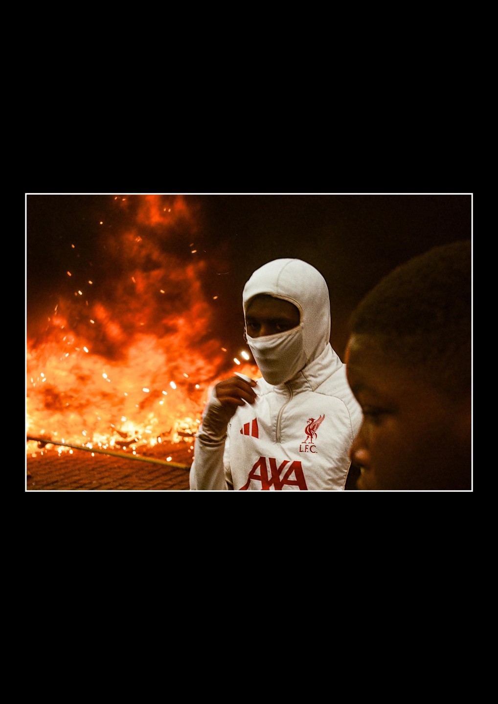 A person in a white balaclava and jacket stands in front of a large fire with sparks flying.