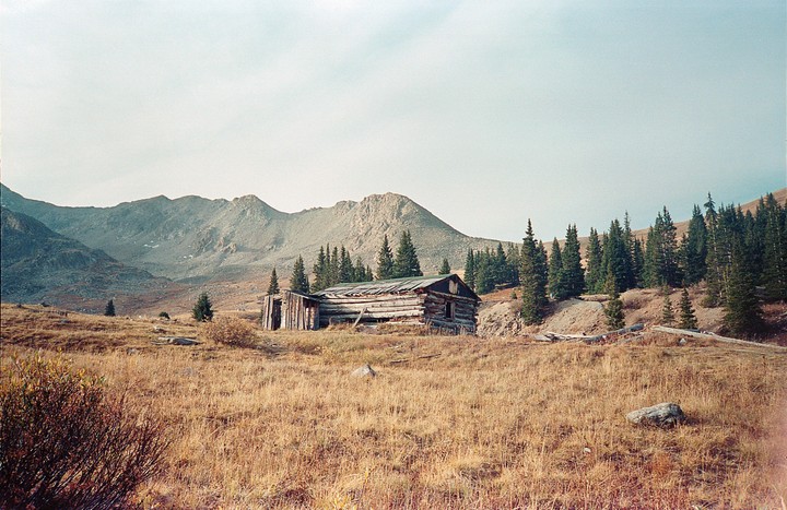 An old wooden cabin sits in a field with mountains and pine trees in the background.