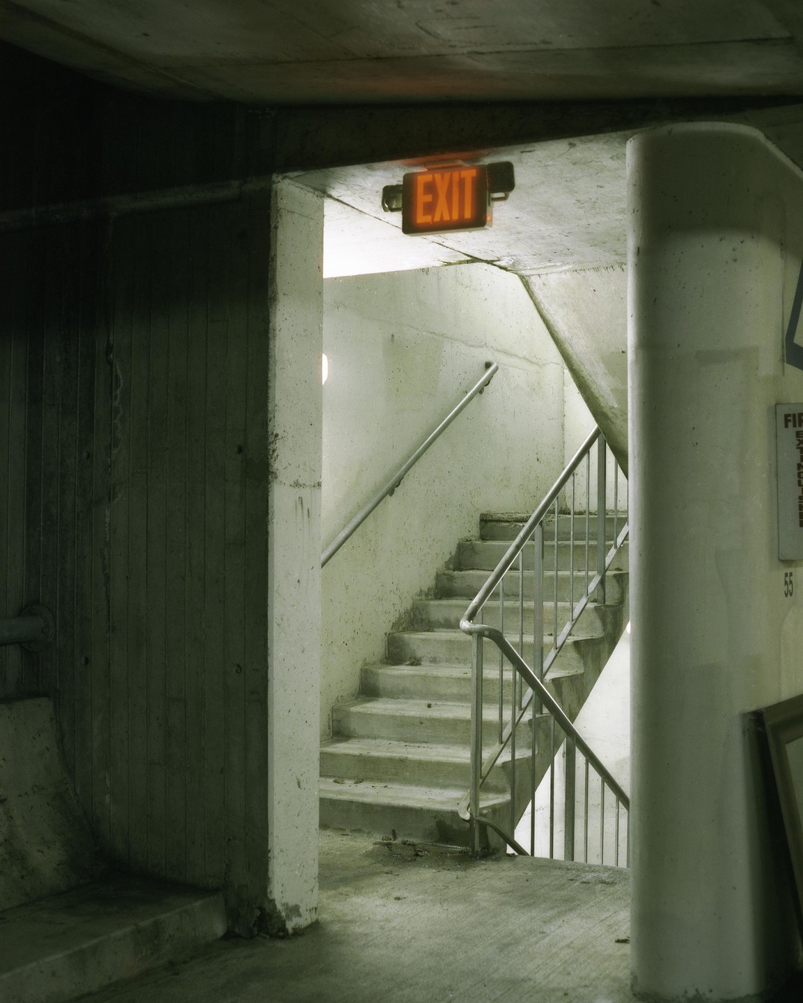 An orange exit sign hangs above a concrete staircase with metal railings in a dimly lit building.