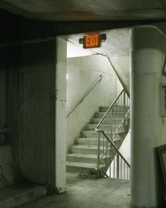 An orange exit sign hangs above a concrete staircase with metal railings in a dimly lit building.