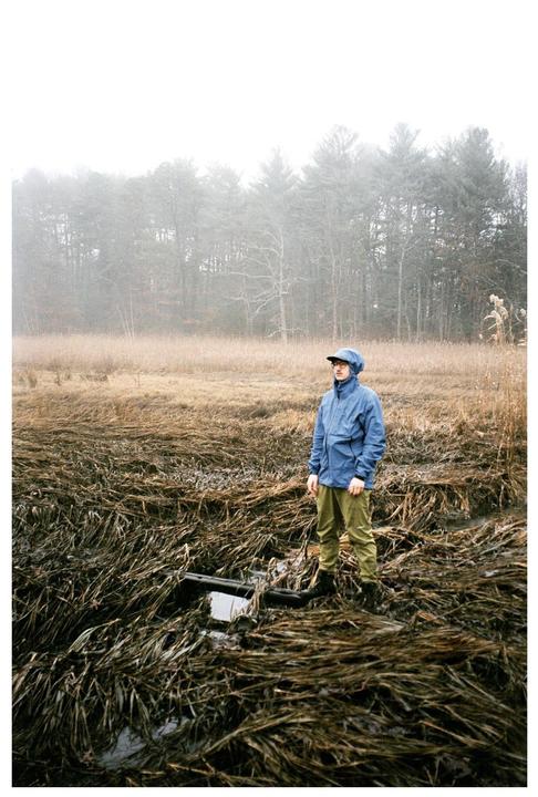 A man in a blue jacket stands in a marsh with tall, dry grass and a forest in the background.