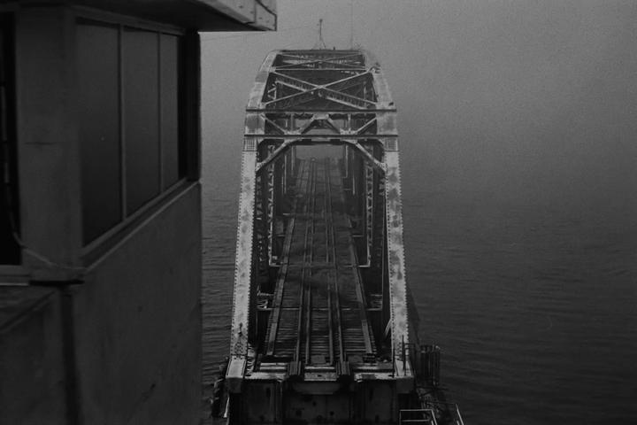 A long, metal railway bridge extends over water into a hazy distance, viewed from a building on the left.