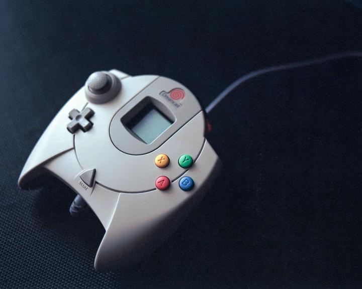 A Dreamcast controller with colorful buttons and a small screen is angled on a dark surface.