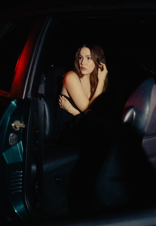 A woman sits in the passenger seat of a car at night, illuminated by a red light.