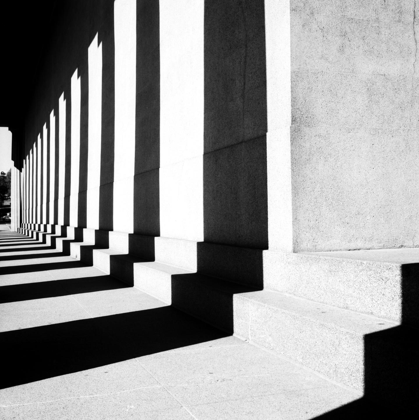 A row of columns casts long shadows on the ground next to a building.
