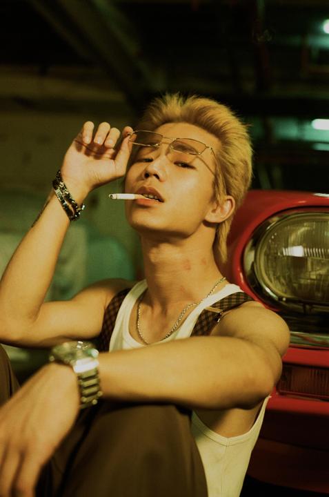 A man with bleached hair and sunglasses holds a cigarette in his mouth while leaning against a red car.