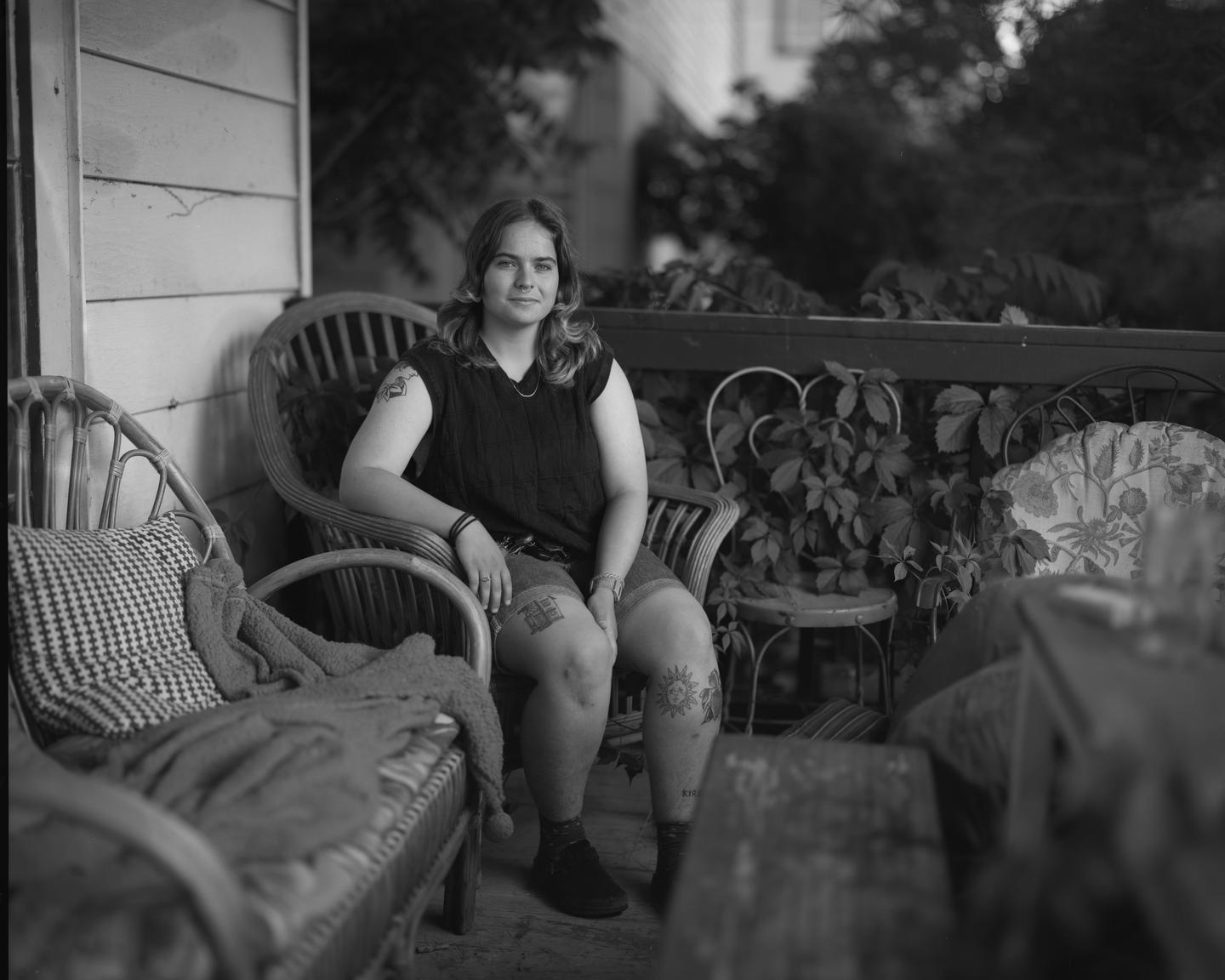 A woman with tattoos sits on a wicker chair on a porch.