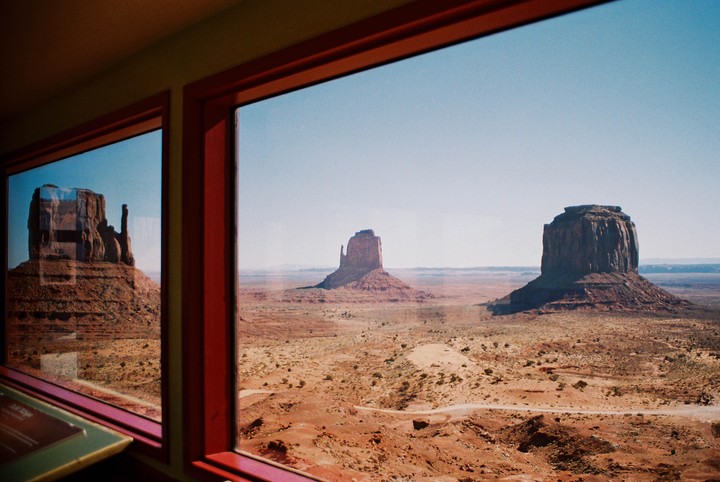 Three large rock formations rise from a vast desert landscape, viewed through a window with red frames.