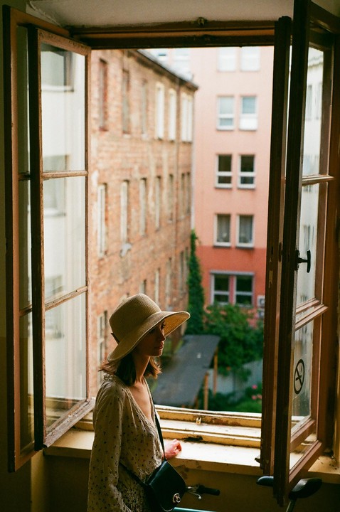 A woman in a straw hat looks out an open window at brick buildings and green trees.