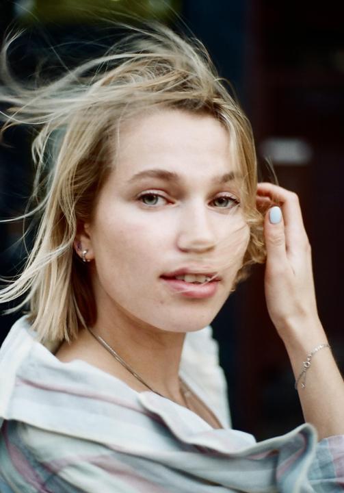 A woman with short blonde hair and a striped shirt looks at the viewer, with her hair blowing in the wind.