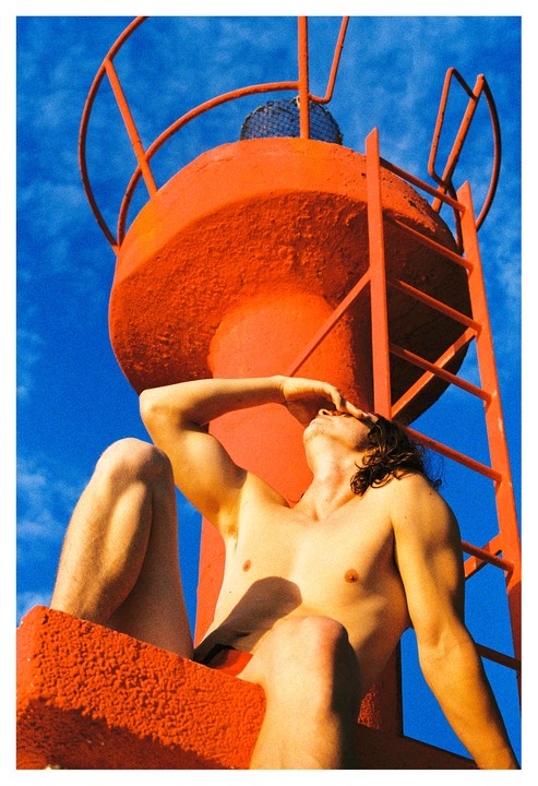 A shirtless man with long hair shields his eyes from the sun while sitting on a large orange buoy with a ladder against a bright blue sky.