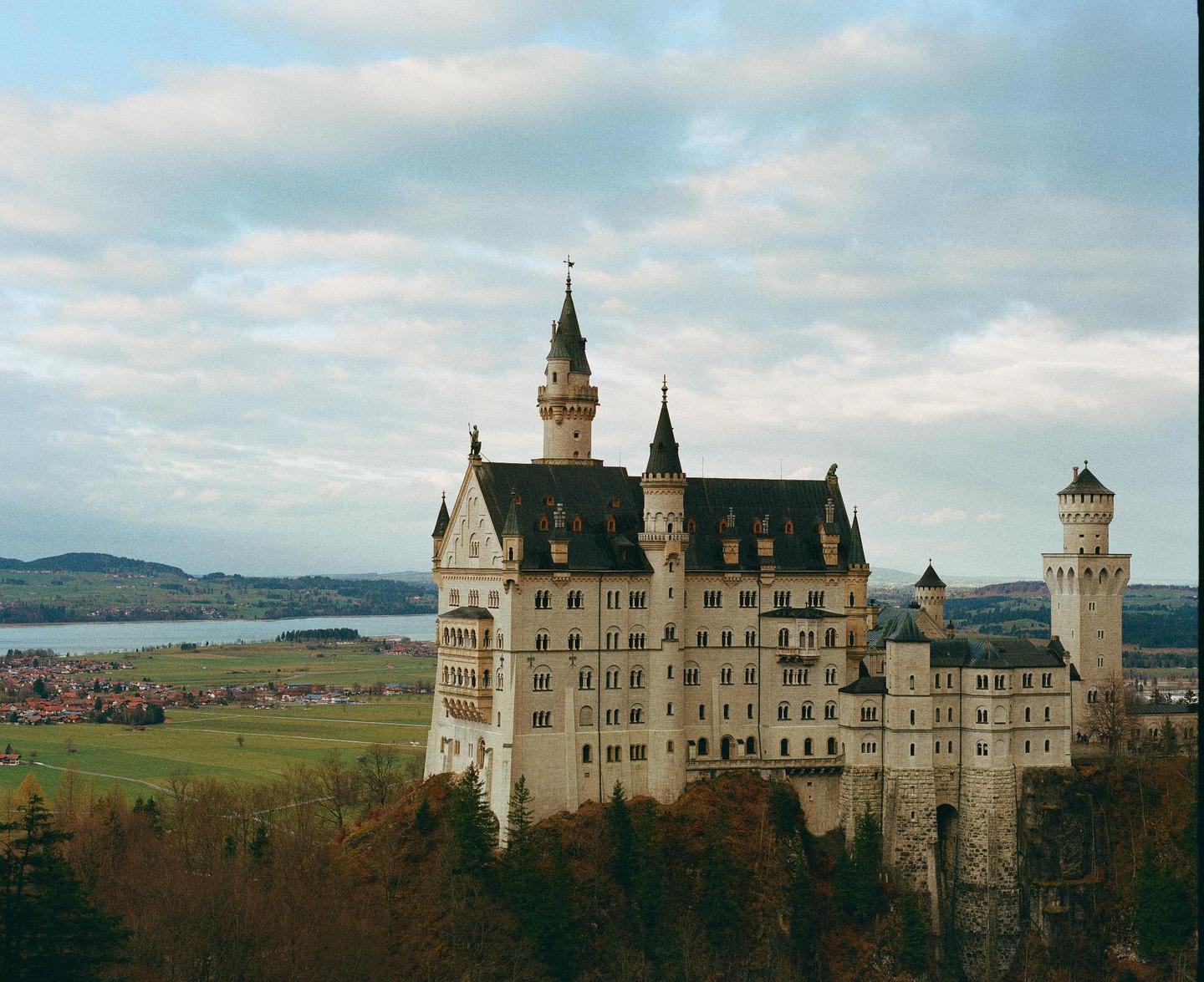 A large castle with multiple towers and spires stands on a rocky outcrop overlooking a valley with a lake and a village.