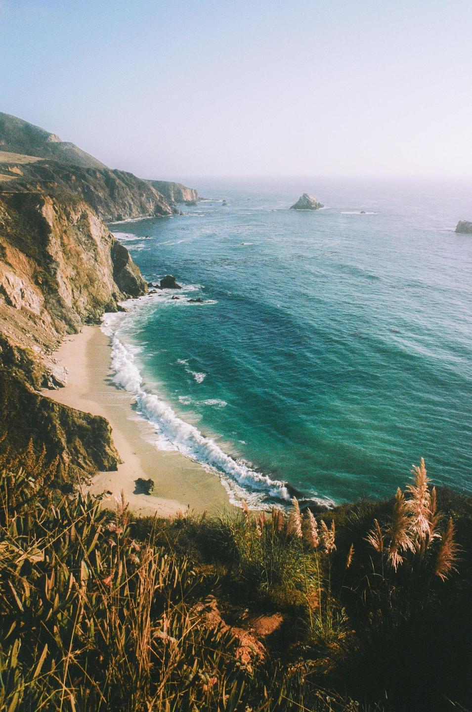 A high-angle landscape shows a sandy beach with waves crashing against a rocky coastline and a vast ocean under a clear sky.