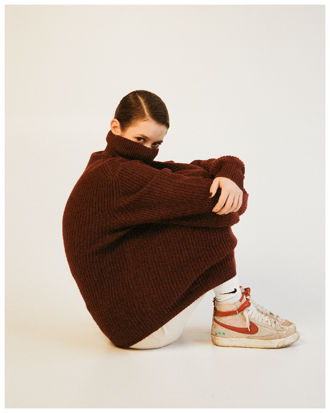 A woman in a dark red turtleneck sweater is sitting with her knees pulled up to her chest, looking directly at the viewer.