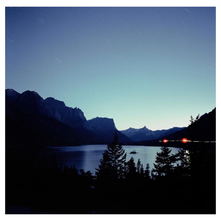 Star trails are visible in the sky above a lake surrounded by mountains and trees at night.