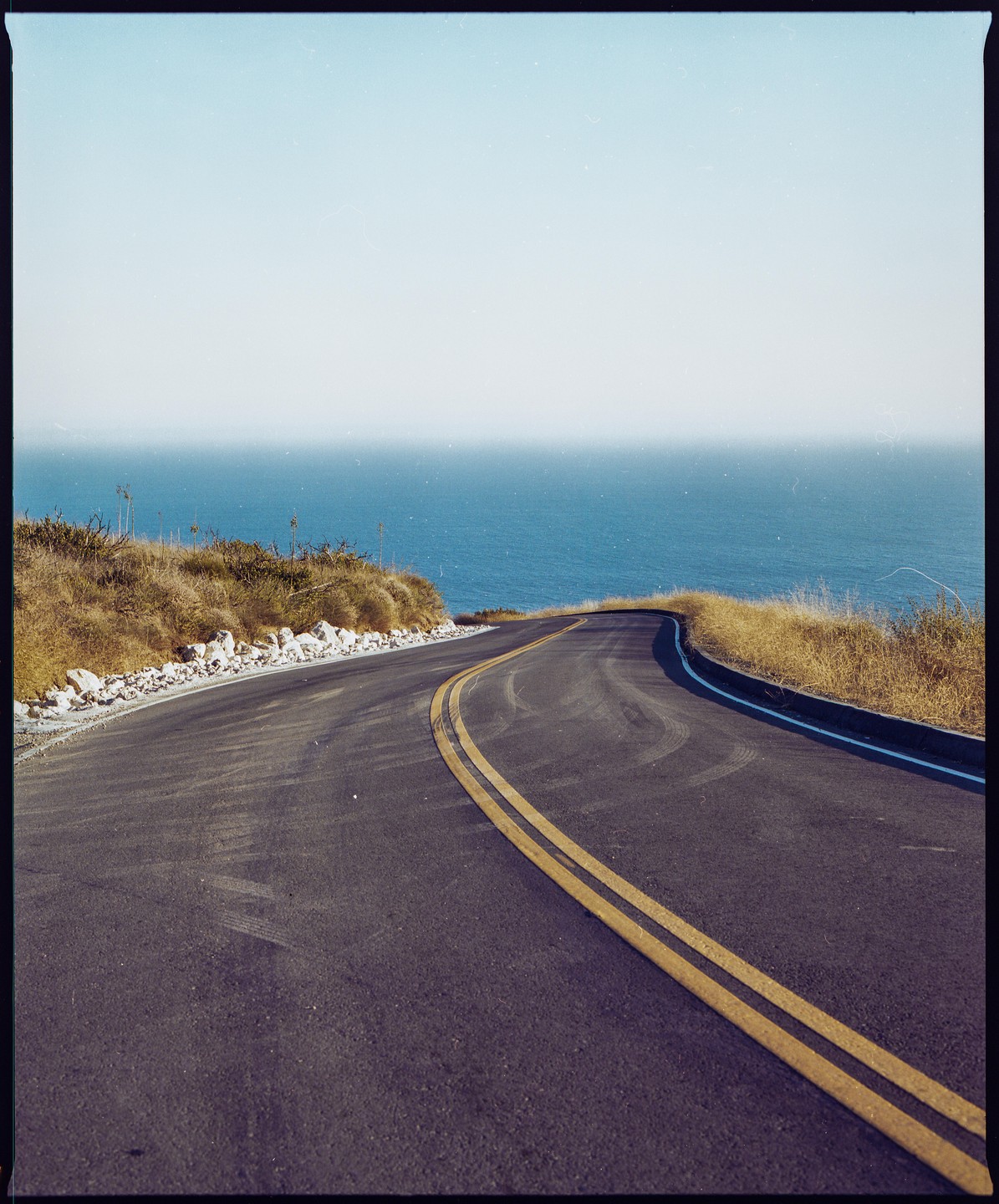 A winding road with double yellow lines curves along a coastline overlooking the ocean.