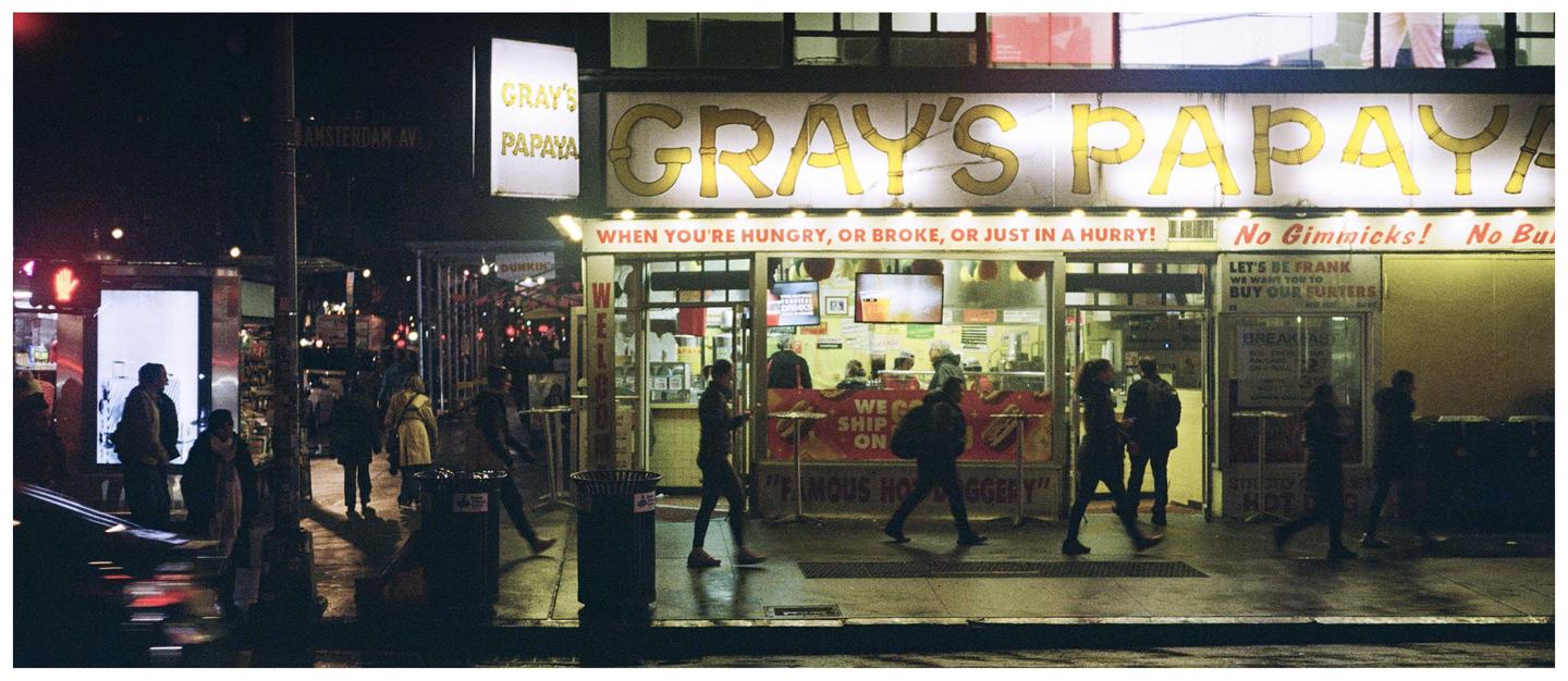 People walk past a Gray's Papaya hot dog stand at night on Amsterdam Avenue.