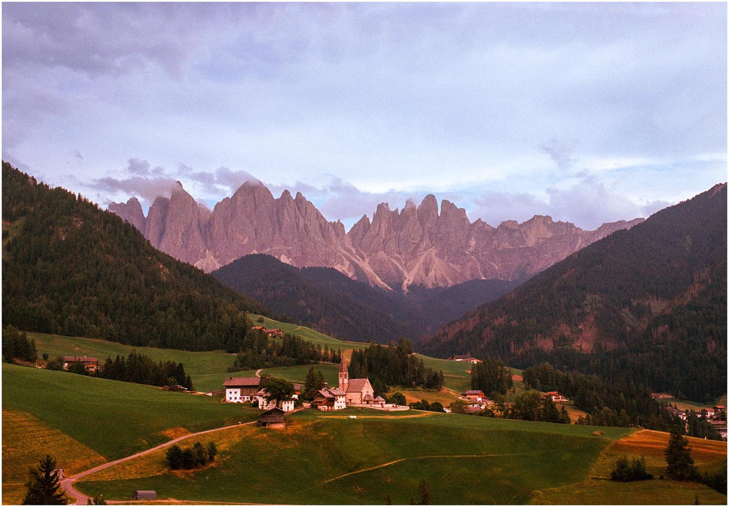 A small village with a church is nestled in a valley surrounded by green hills and towering, jagged mountains under a cloudy sky.