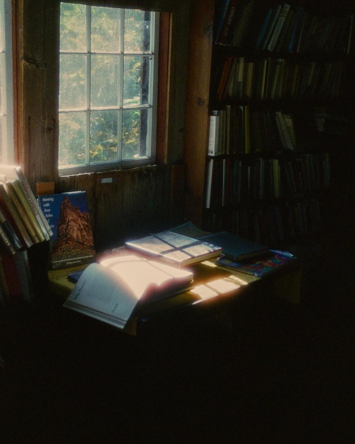 Sunlight streams through a window onto books on a table and a bookshelf in a dimly lit room.