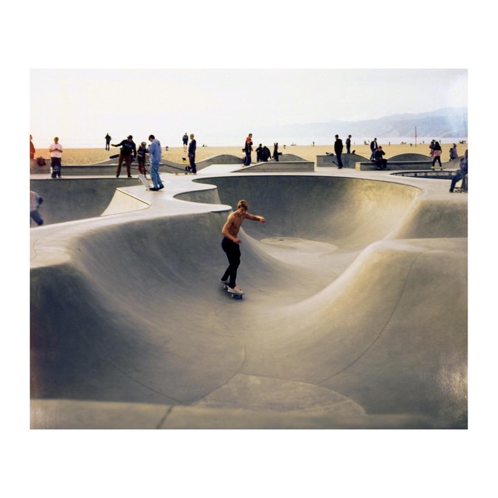 A shirtless man skates in a concrete skatepark with a beach and mountains in the background.