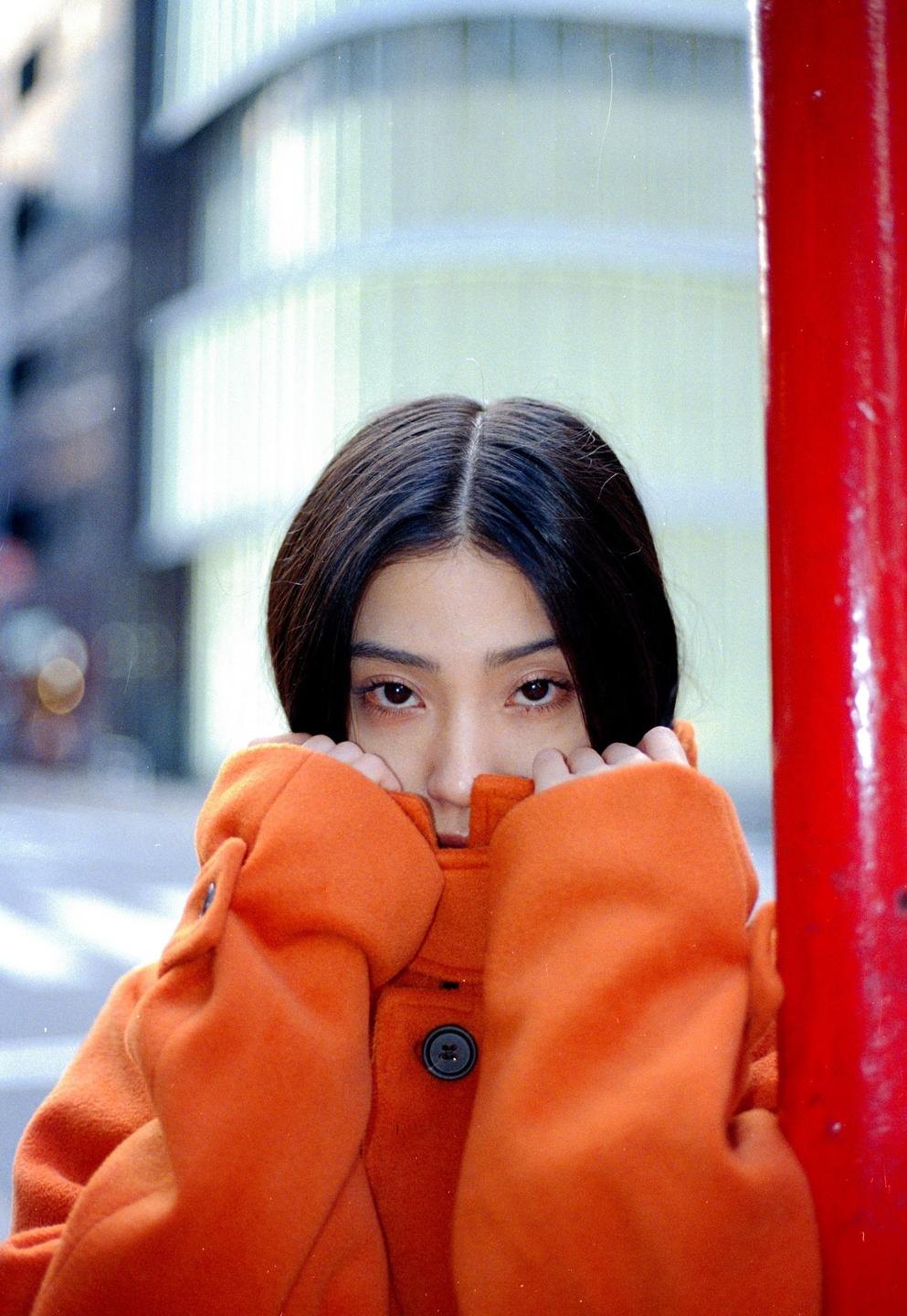 A woman in an orange coat covers her face with her hands while looking directly at the viewer.