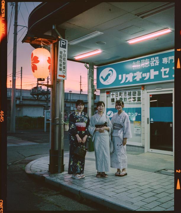 Three women in kimonos stand on a sidewalk next to a building with a paper lantern hanging from a pole.