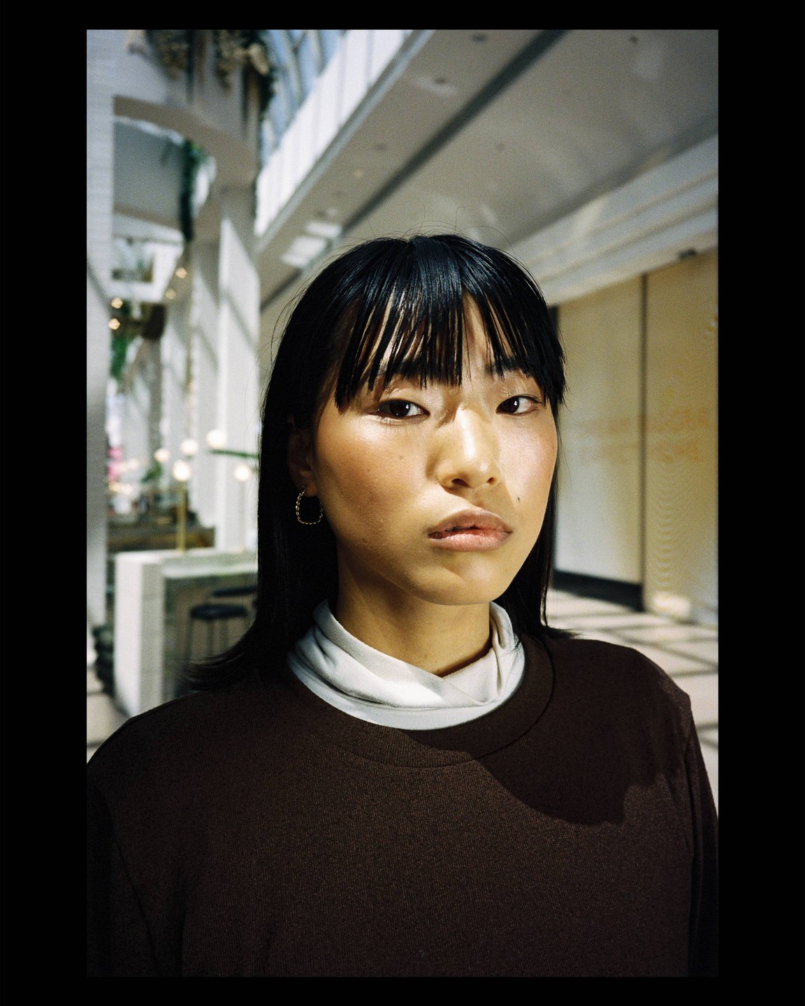 A woman with dark hair and bangs wears a brown shirt and a white turtleneck.
