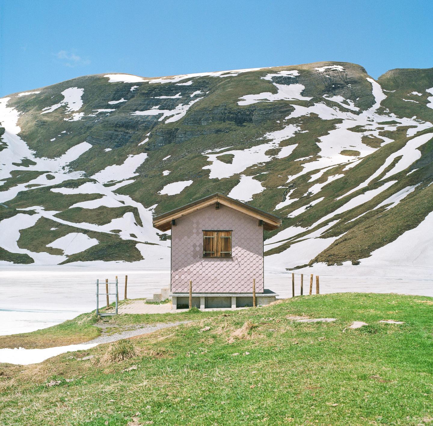 A small pink house with a brown roof sits on a grassy hill next to a frozen lake, with a snow-covered mountain in the background.