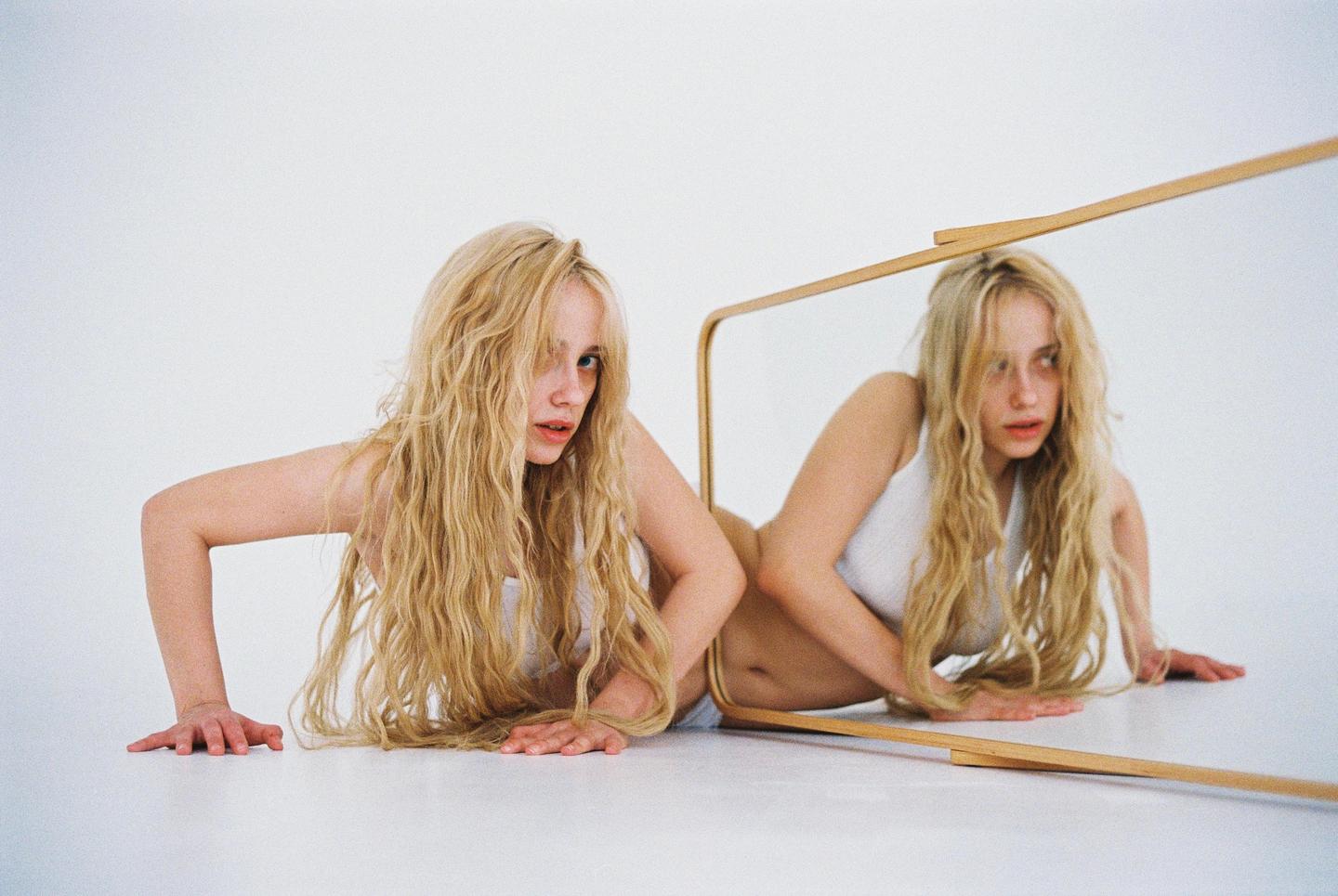 A woman with long blonde hair is reflected in a mirror while lying on a white surface.