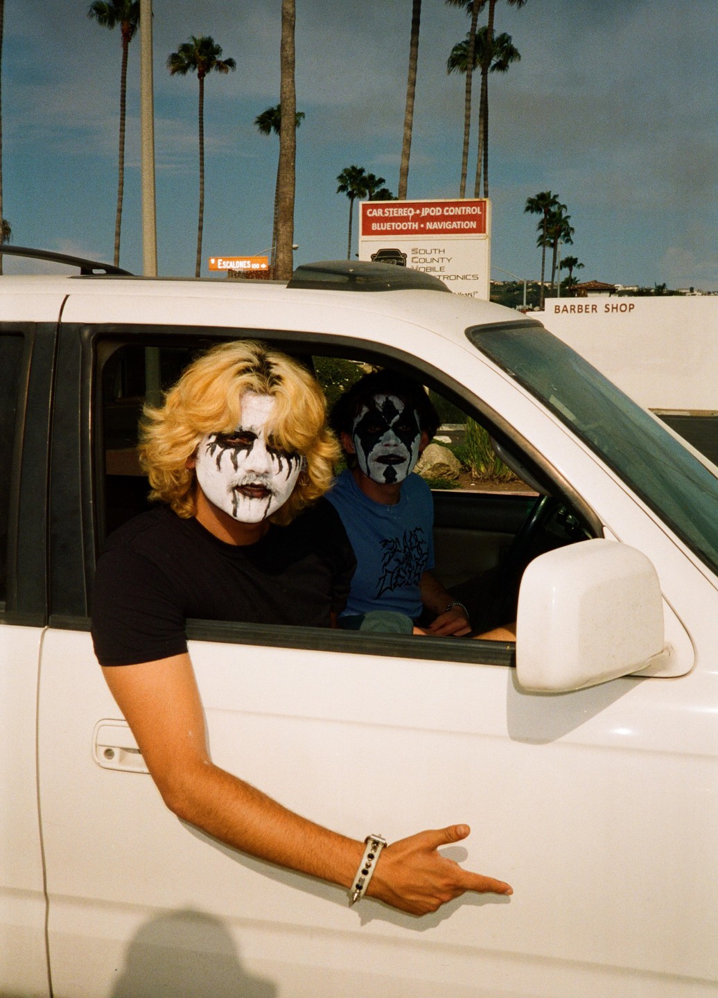 Two men with face paint are in a white car with palm trees and signs in the background.