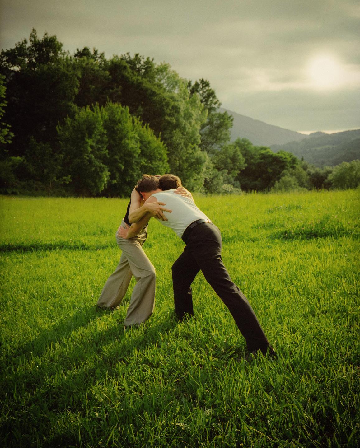 Two men wrestle in a grassy field with trees and mountains in the background under a bright sun.