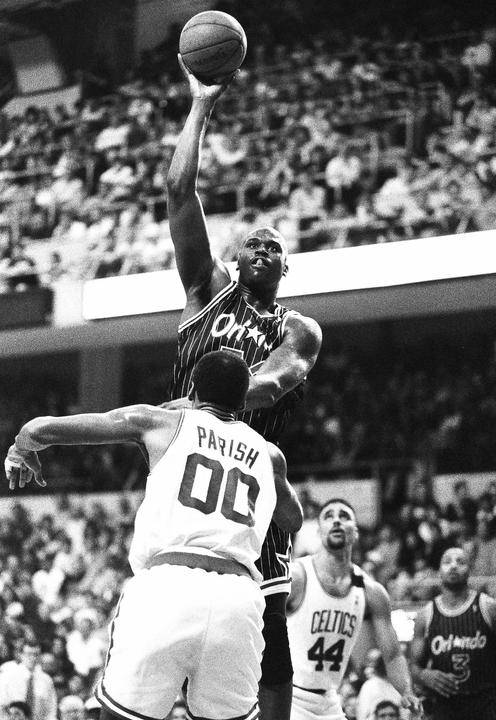 A basketball player in a striped jersey shoots a basketball over an opponent in a white jersey, with other players and spectators in the background.
