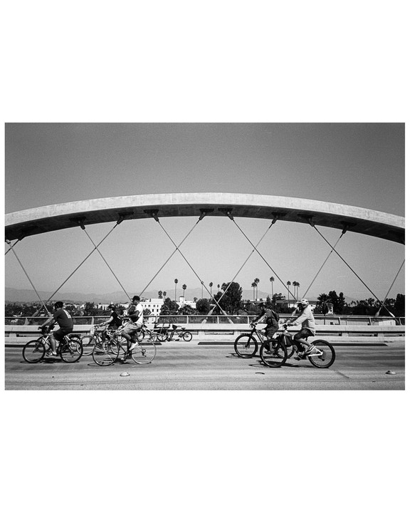 Several people ride bicycles across a bridge with palm trees and buildings in the background.