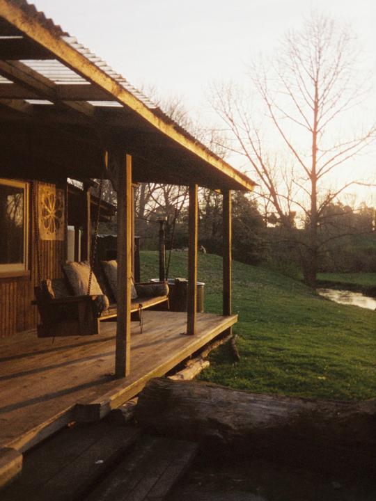 A wooden porch with a swing overlooks a grassy bank and a river at sunset.