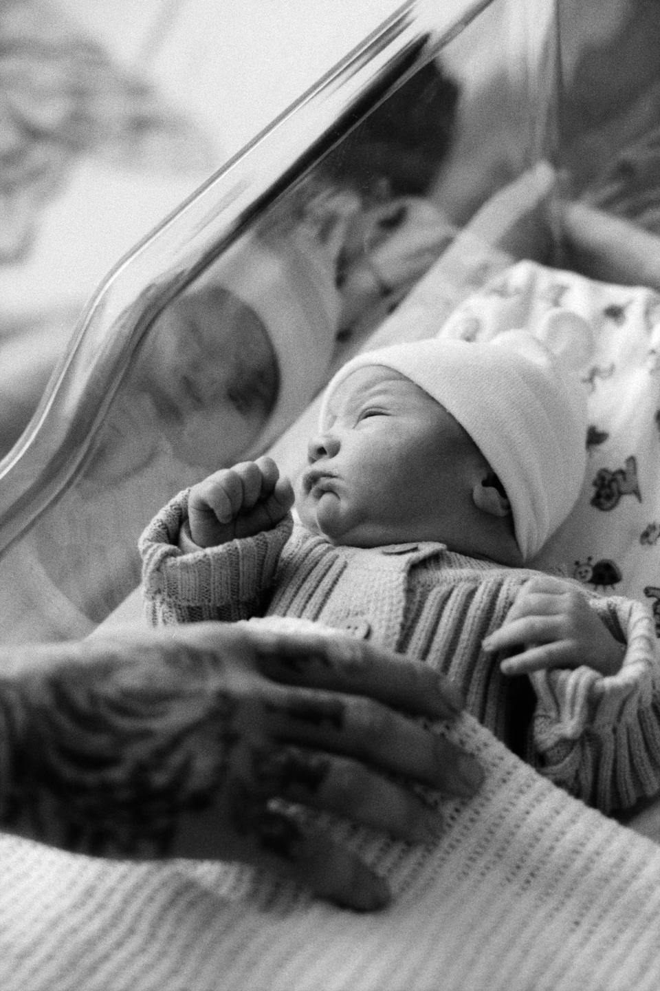 A newborn baby in a knitted sweater and hat lies in a hospital bassinet with a hand resting on the blanket.