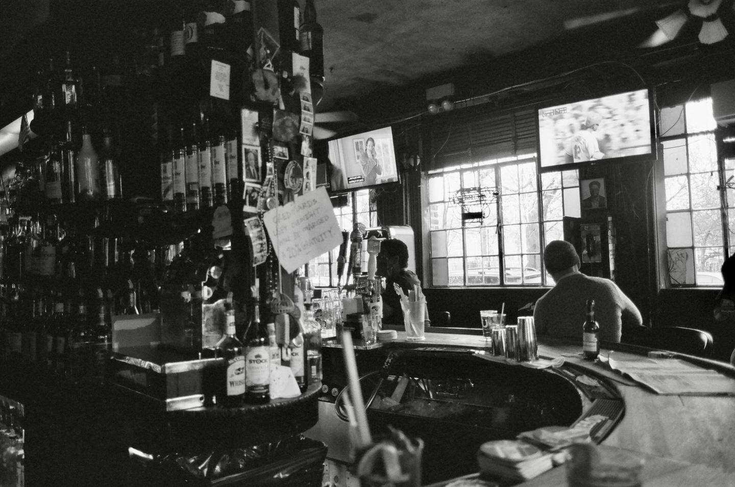 Two men sit at a bar with bottles of alcohol on shelves behind them and televisions above the windows.
