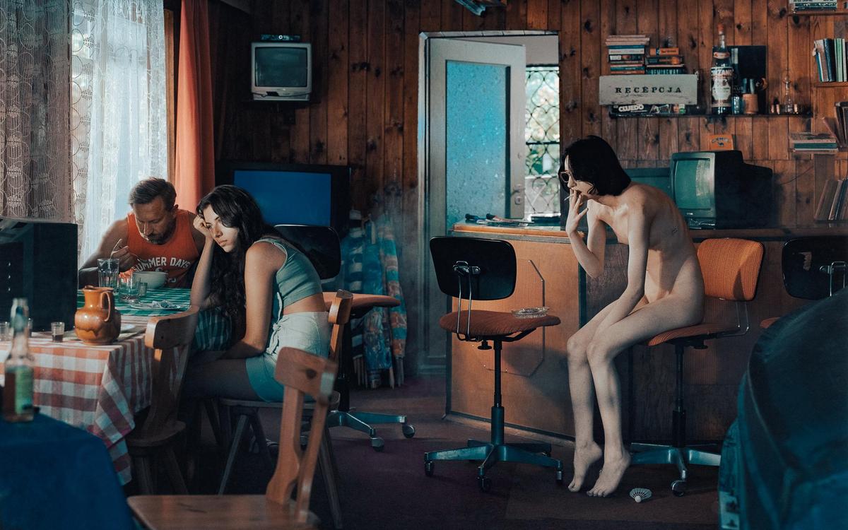 A man and a woman sit at a table while another woman sits nude on a chair smoking a cigarette in a wood-paneled room.